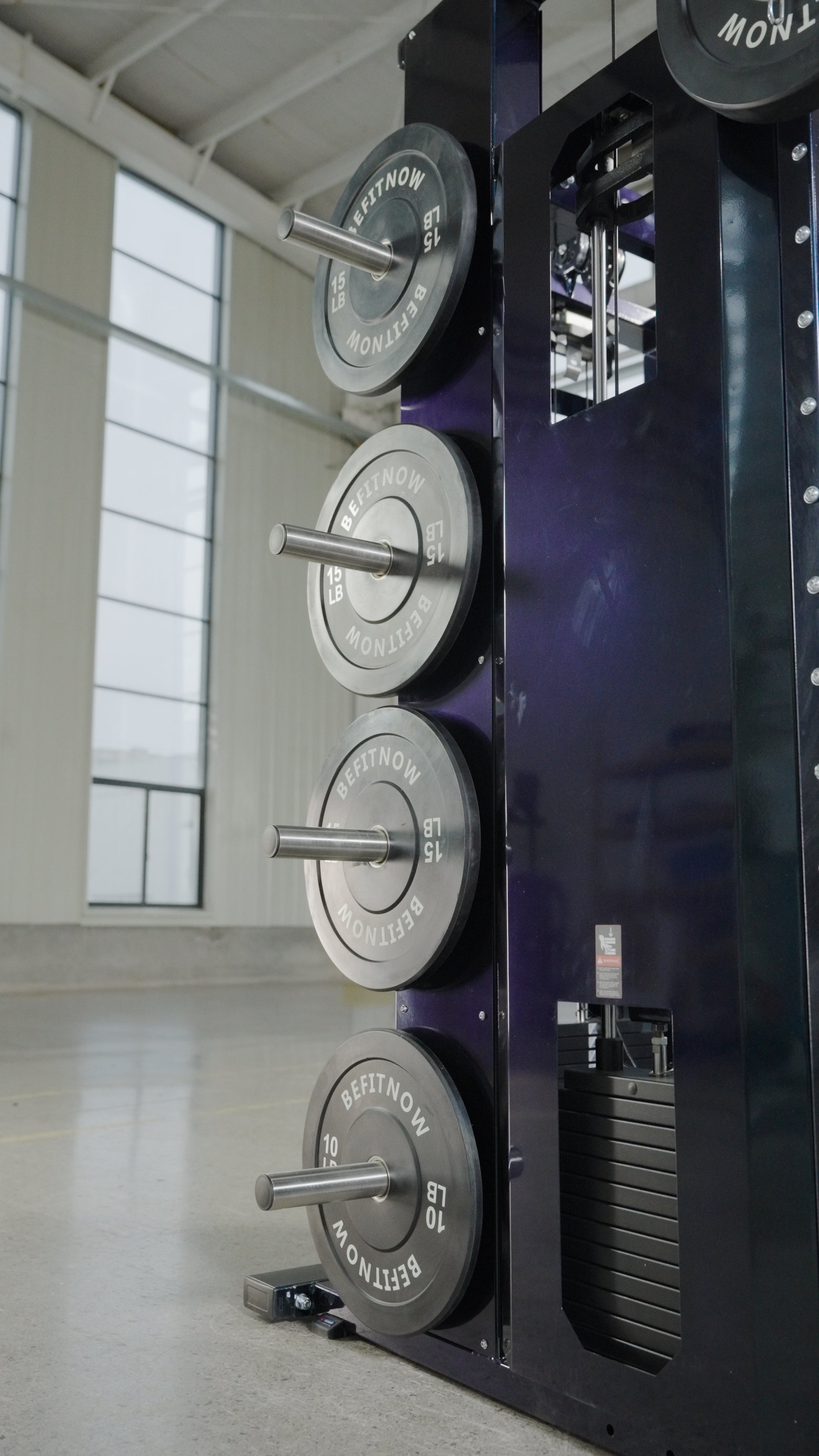 Four BefitNow Canada The Professor weight plates are racked on a functional trainer in a spacious, well-lit gym featuring large windows and concrete floors.