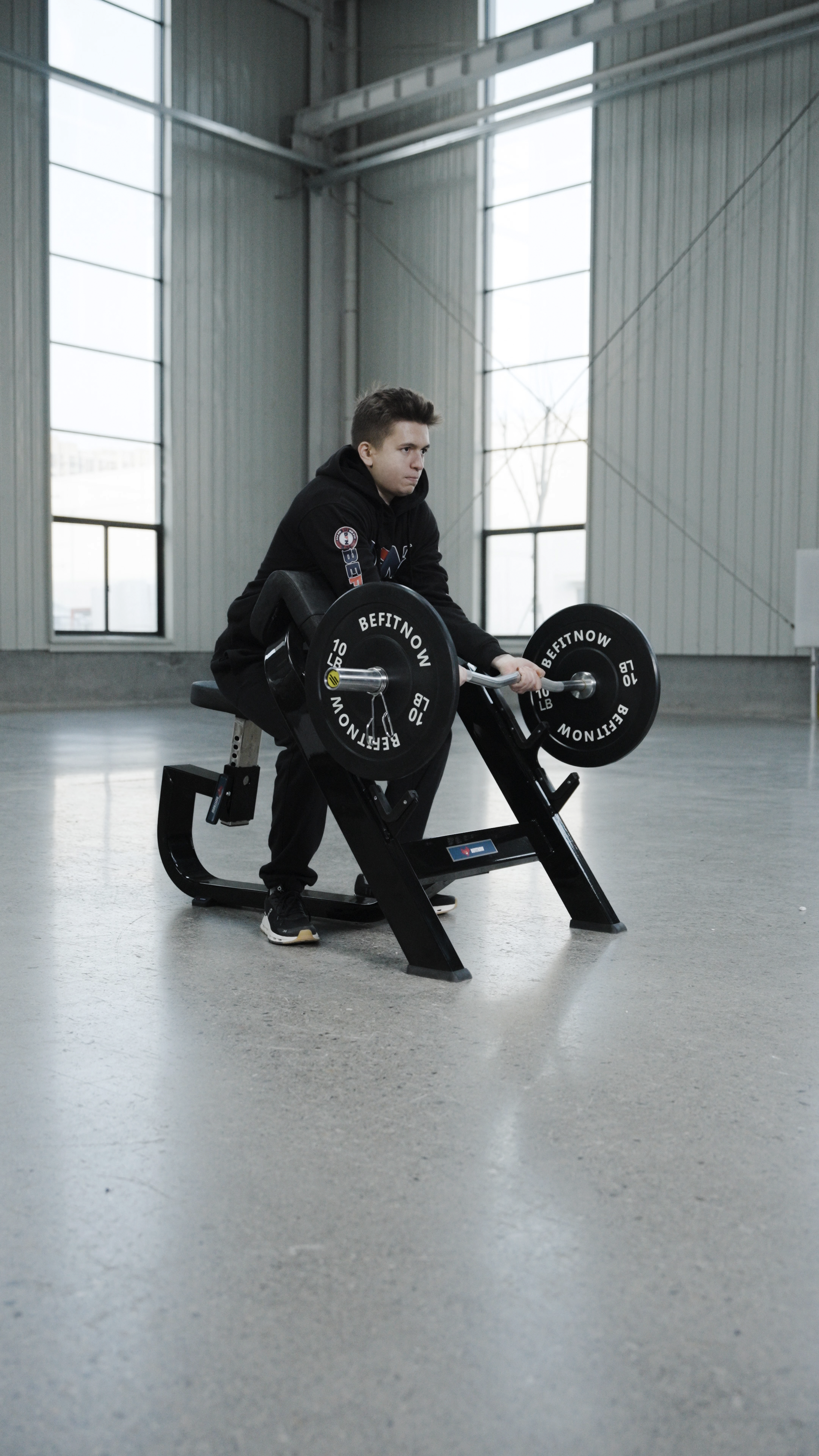 A person in a black hoodie and pants sits indoors on the BefitNow Canada Seated Preacher Curl Bench, gripping its handles. The spacious room with large windows and light gray walls easily fits the 53"x30"x37" bench.
