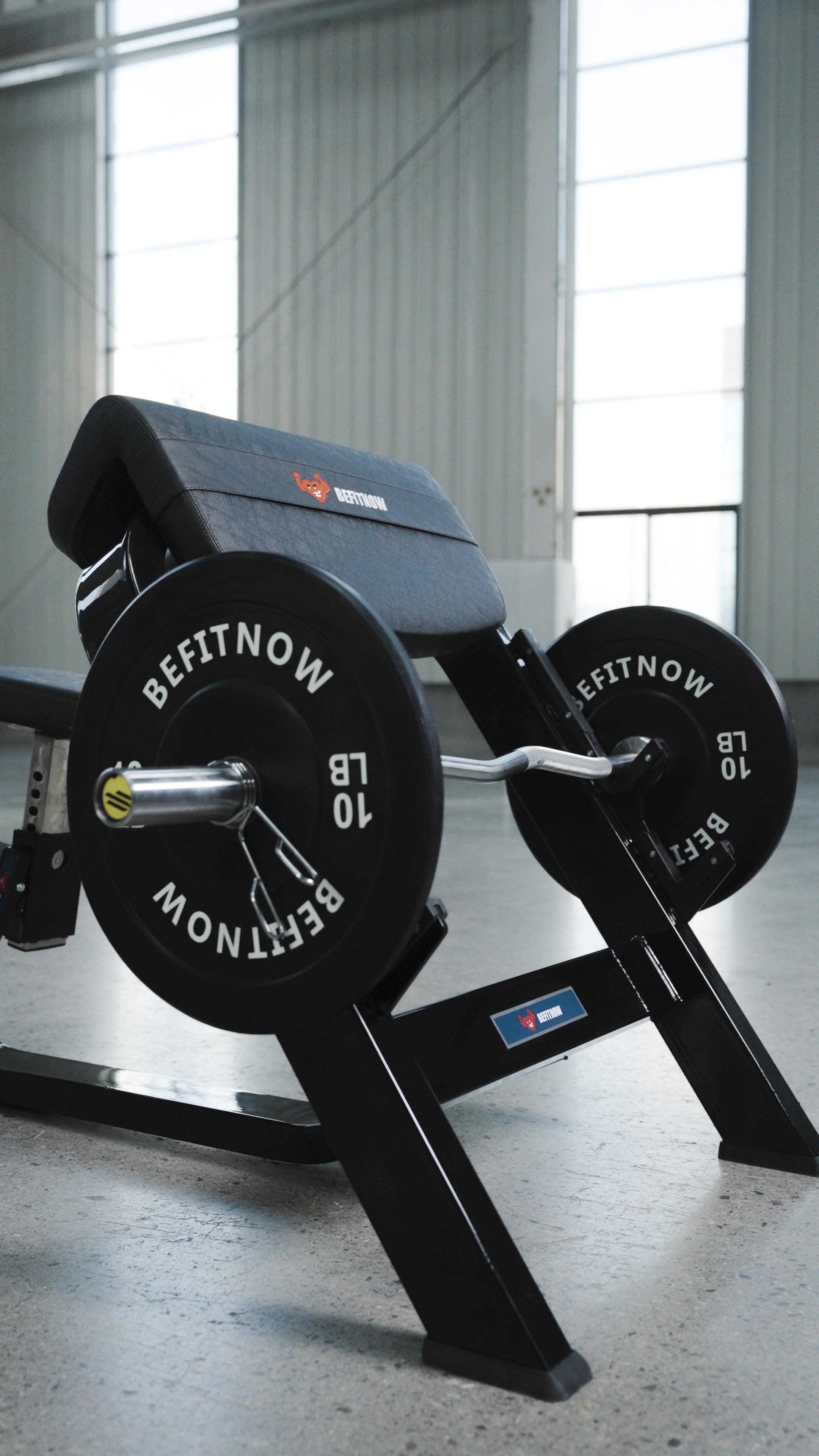 A close-up of the BefitNow Canada Seated Preacher Curl Bench with a loaded barbell and shiny weight plates in a bright gym with large windows and concrete floors; bench measures 53"x30"x37".