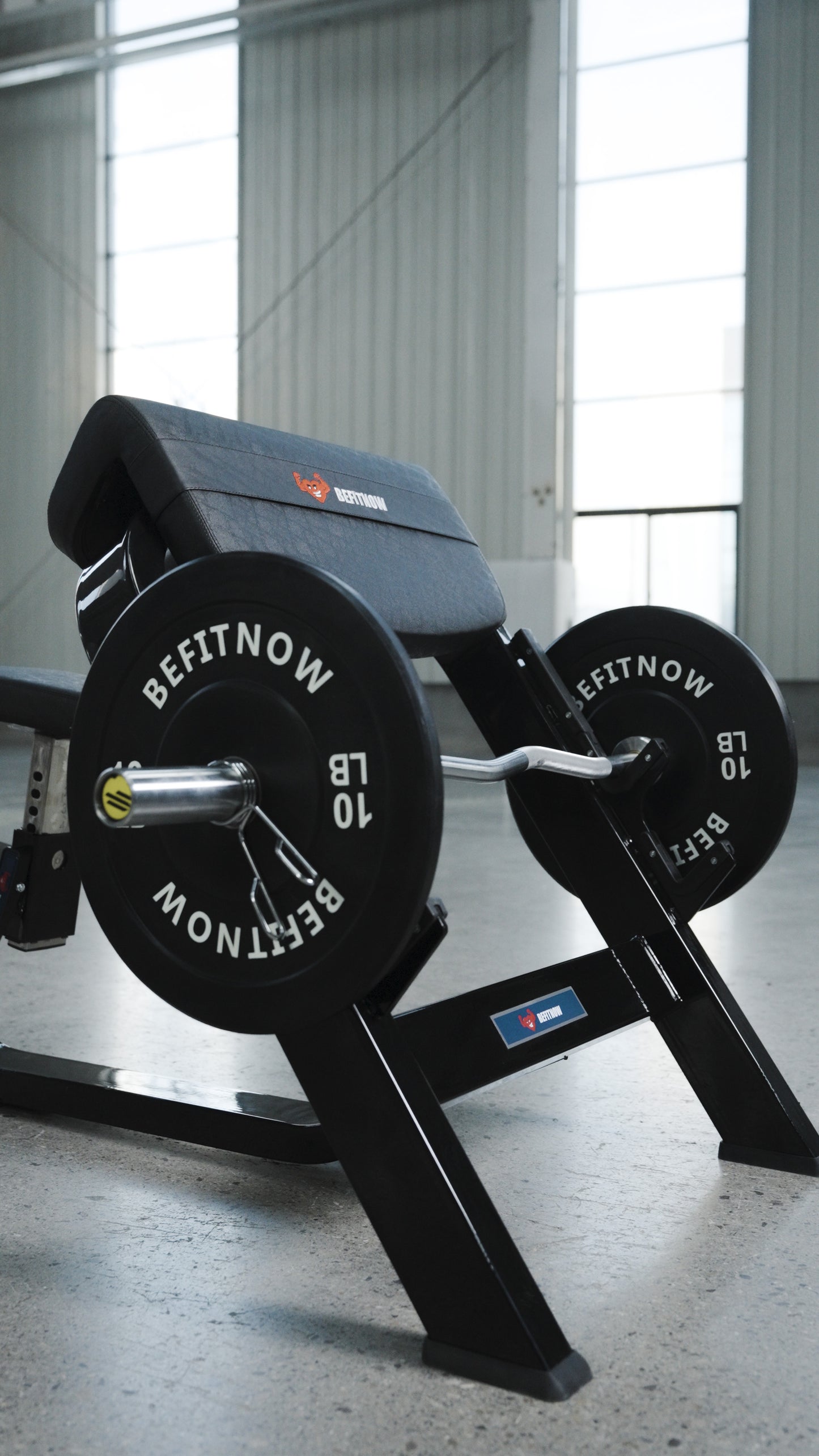 A close-up of the BefitNow Canada Seated Preacher Curl Bench with a loaded barbell and shiny weight plates in a bright gym with large windows and concrete floors; bench measures 53"x30"x37".