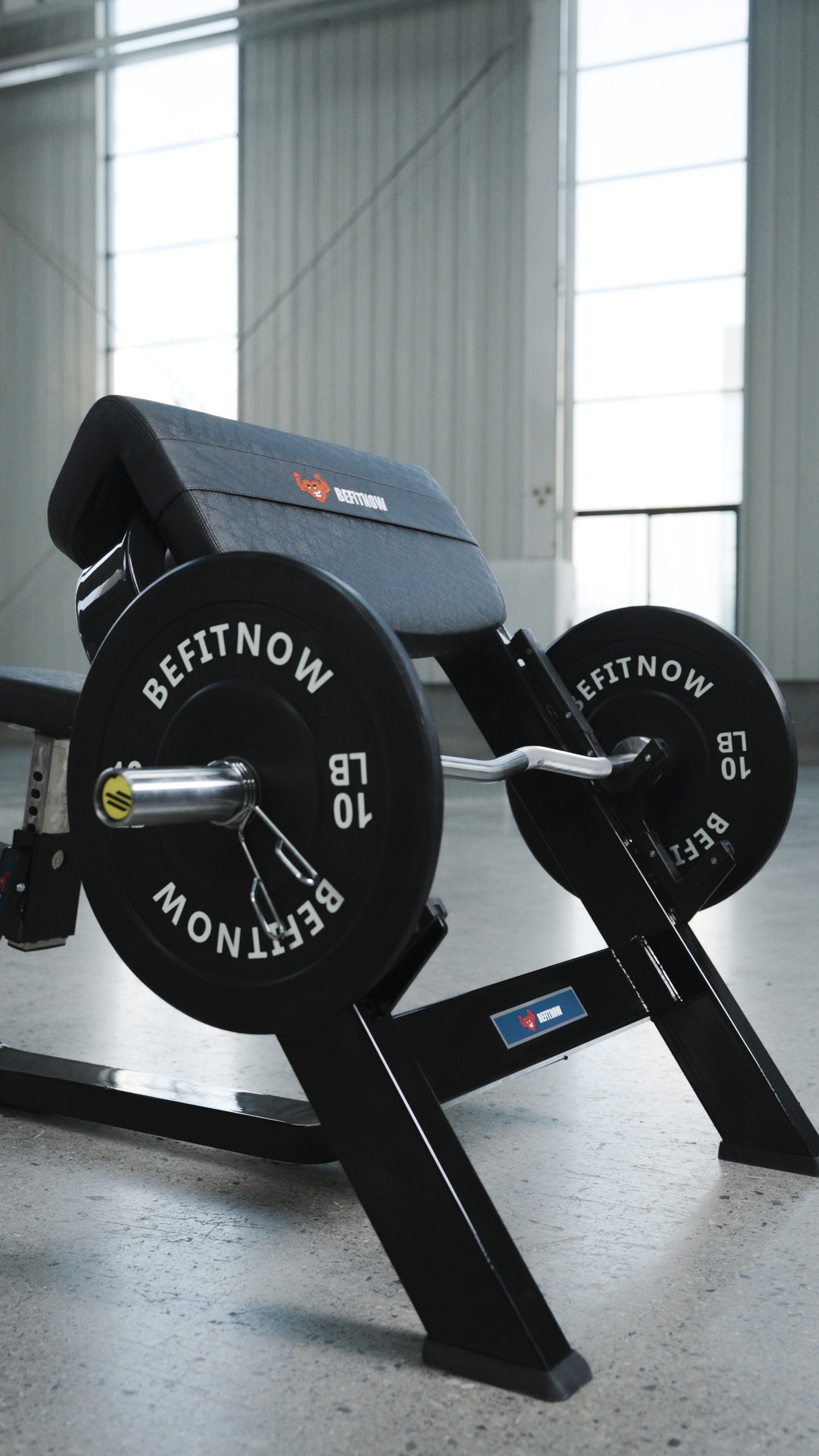 A close-up of the BefitNow Canada Seated Preacher Curl Bench with a loaded barbell and shiny weight plates in a bright gym with large windows and concrete floors; bench measures 53"x30"x37".