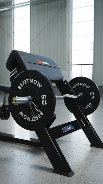 A close-up of the BefitNow Canada Seated Preacher Curl Bench with a loaded barbell and shiny weight plates in a bright gym with large windows and concrete floors; bench measures 53"x30"x37".