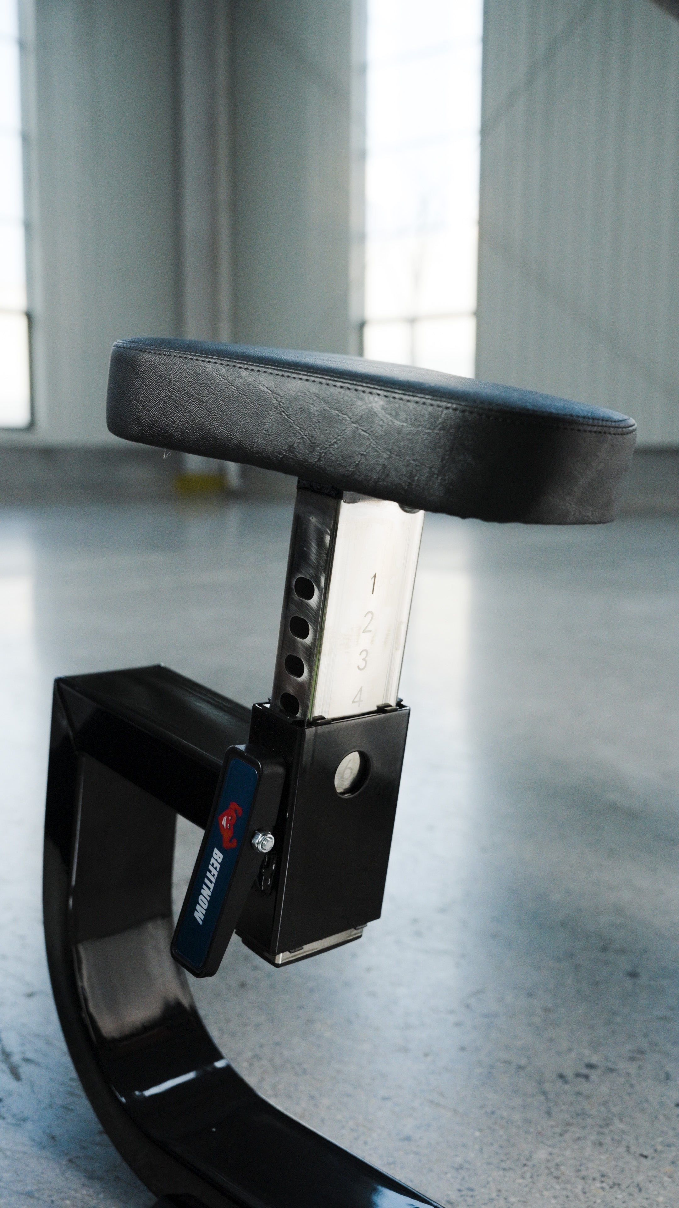 A close-up of the BefitNow Canada Seated Preacher Curl Bench shows its sturdy black padded, adjustable seat with numbered heights on a metal post (53"x30"x37"), placed indoors on concrete with large windows in the background.