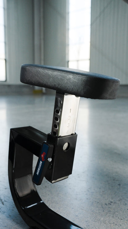 A close-up of the BefitNow Canada Seated Preacher Curl Bench shows its sturdy black padded, adjustable seat with numbered heights on a metal post (53"x30"x37"), placed indoors on concrete with large windows in the background.