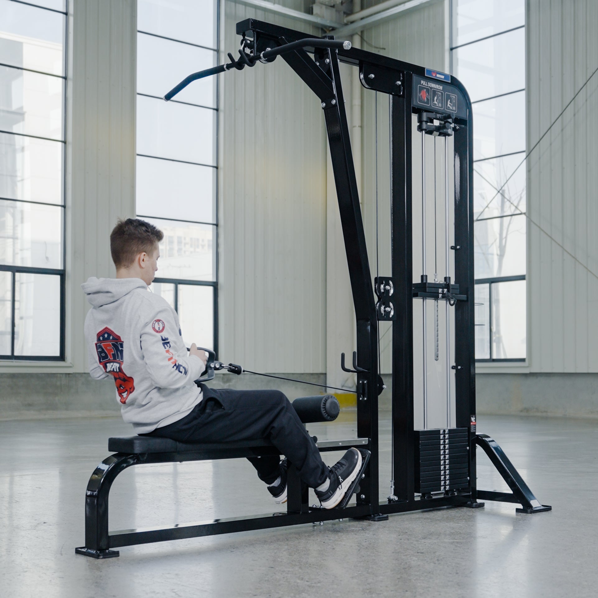 A person in a gray hoodie and black pants uses the BefitNow Canada Lat Pulldown/Low Row machine in a bright gym with large windows, concrete floors, and commercial gym equipment.
