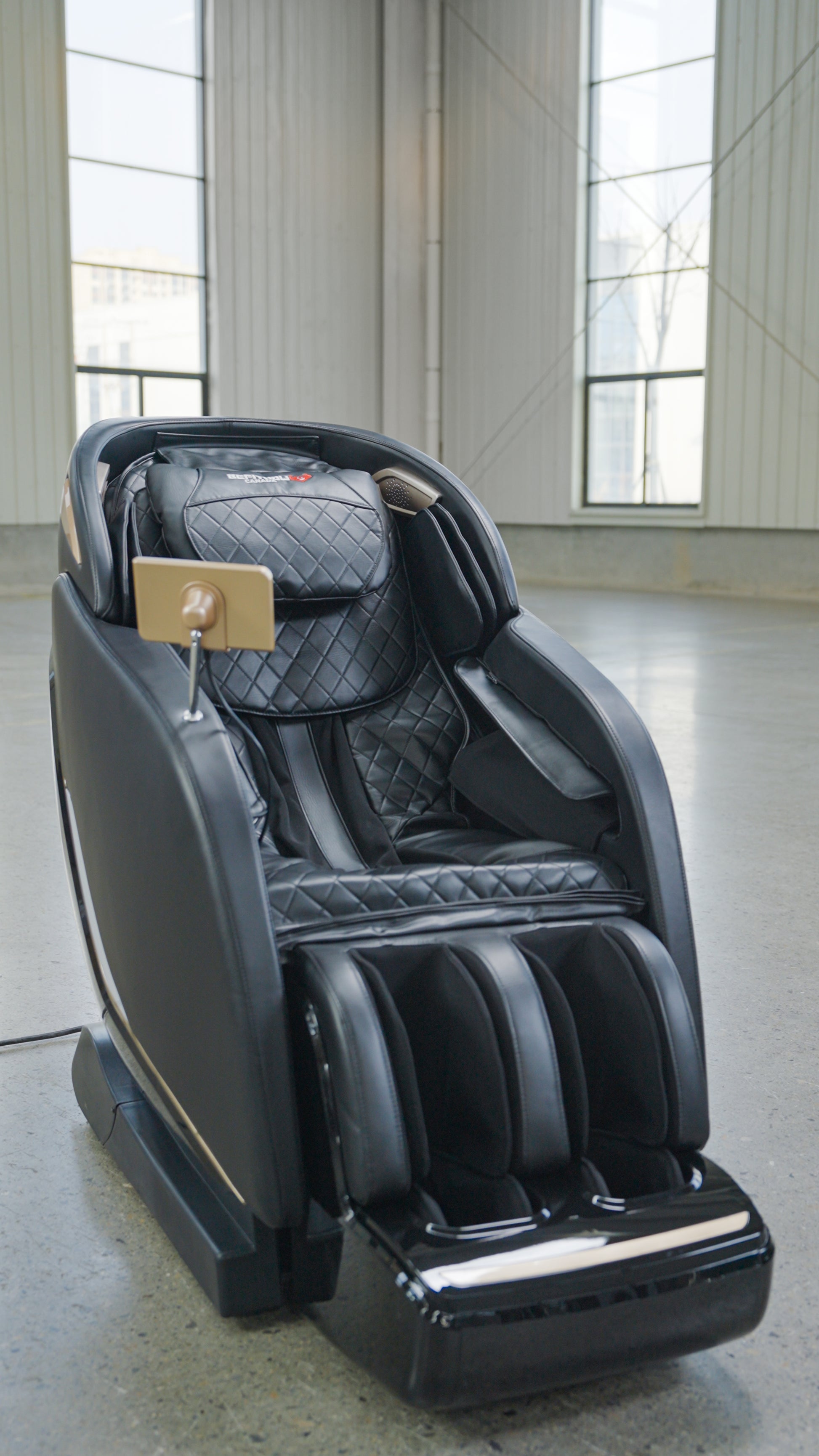 The BefitNow Canada SL Full Body Commercial Massage Chair, featuring quilted black upholstery and a touchpad control on the armrest, offers a relaxing full-body massage in spacious rooms with large windows and concrete floors.