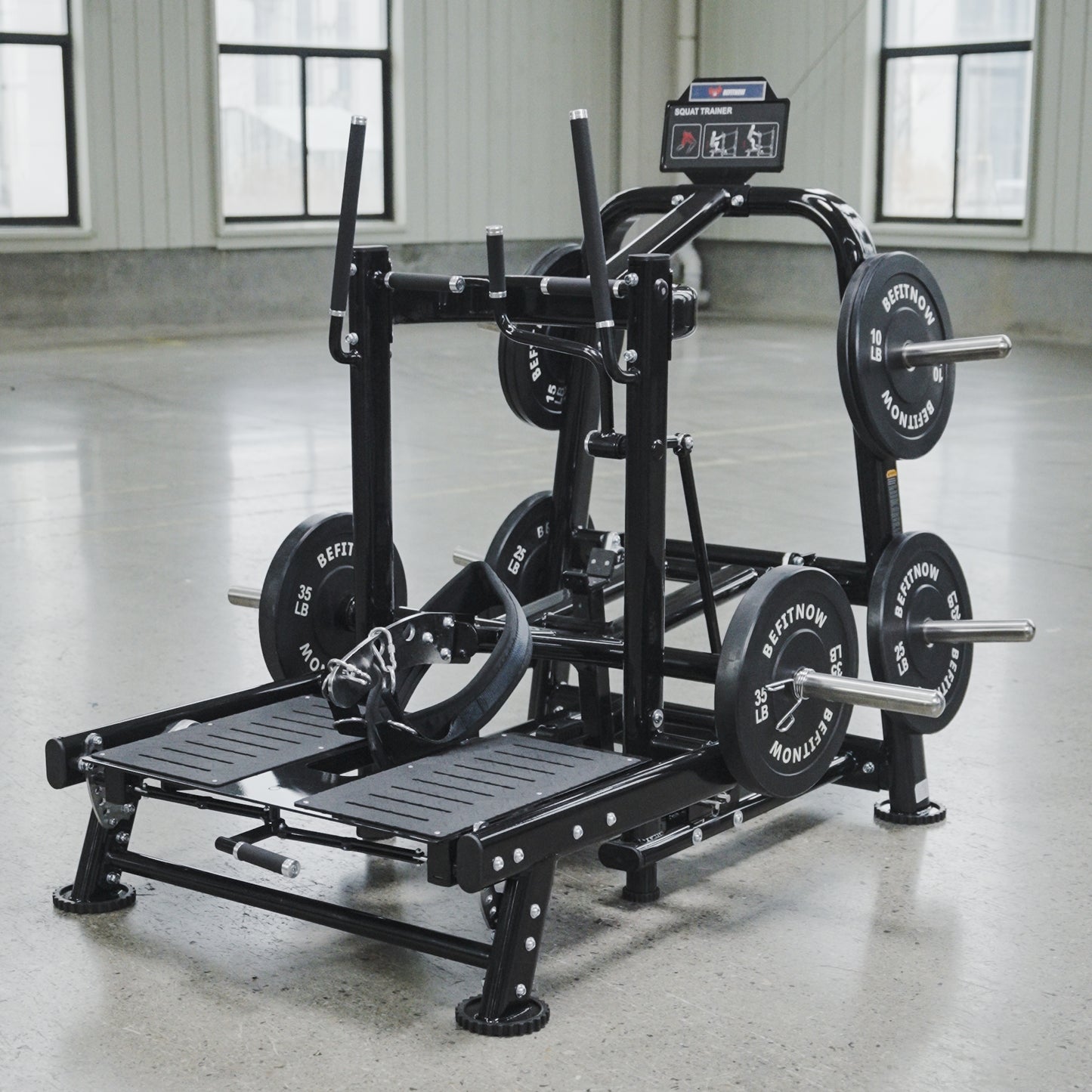 The BefitNow Canada Commercial Belt Squat Trainer, a black multifunctional fitness machine with weights and foot platforms, stands in a spacious gym with concrete floors and large windows.