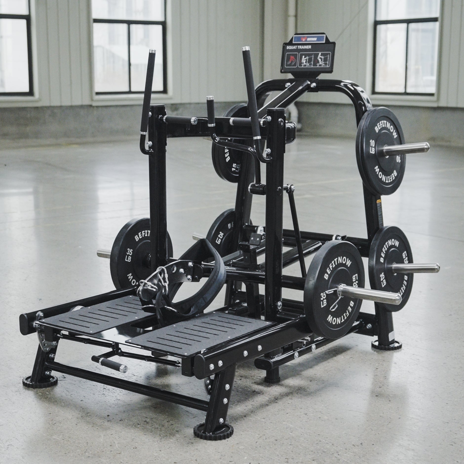 The BefitNow Canada Commercial Belt Squat Trainer, a black multifunctional fitness machine with weights and foot platforms, stands in a spacious gym with concrete floors and large windows.