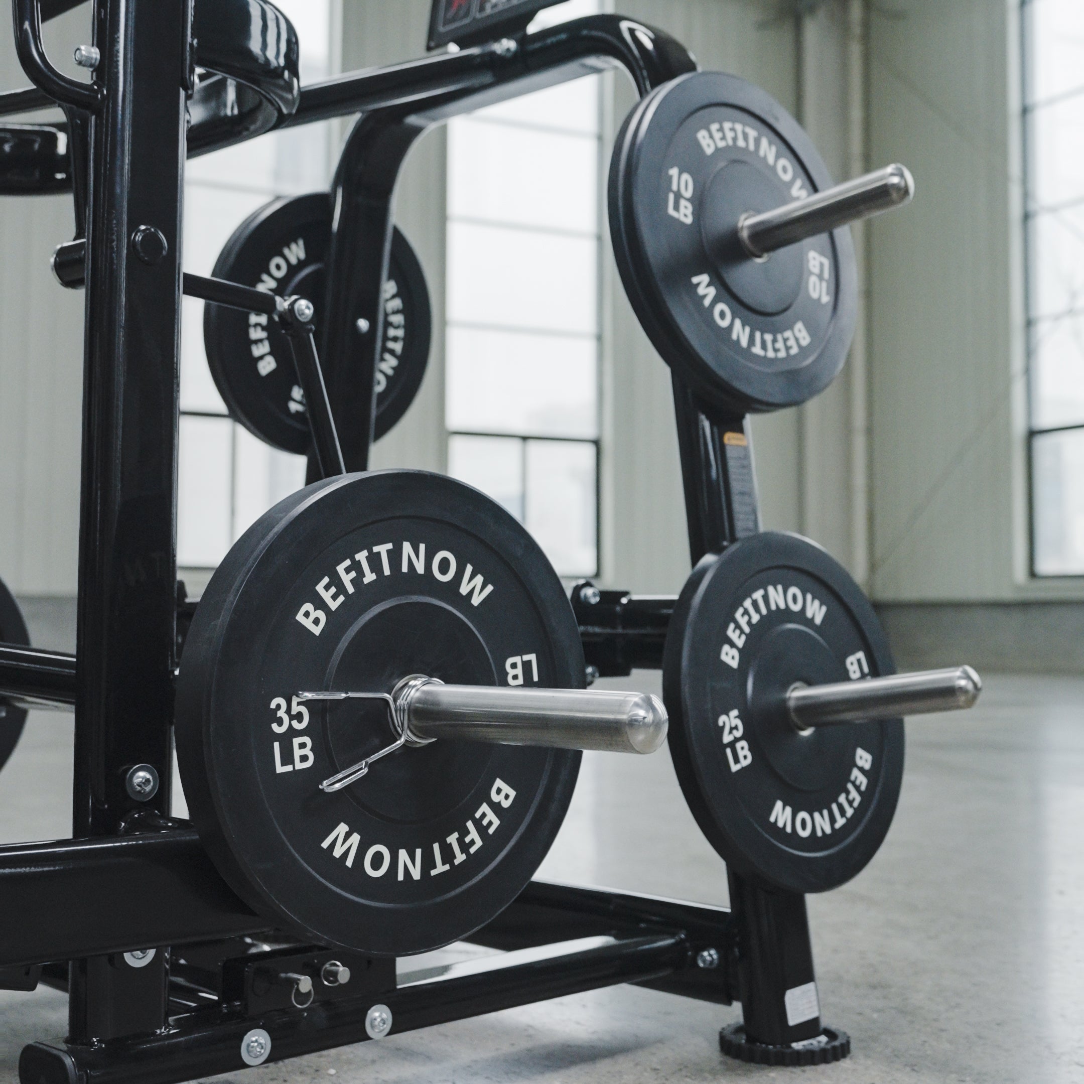 A close-up of the BefitNow Canada Commercial Belt Squat Trainer showing black weight plates (10 LB, 25 LB, 35 LB) on racks in a bright, spacious gym with large windows.