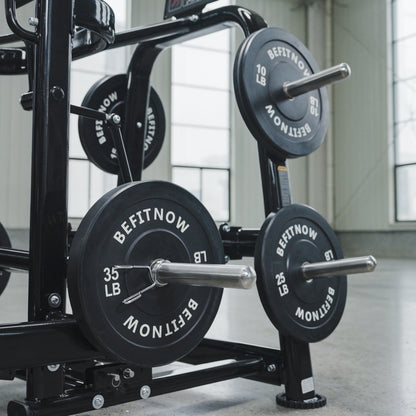 A close-up of the BefitNow Canada Commercial Belt Squat Trainer showing black weight plates (10 LB, 25 LB, 35 LB) on racks in a bright, spacious gym with large windows.