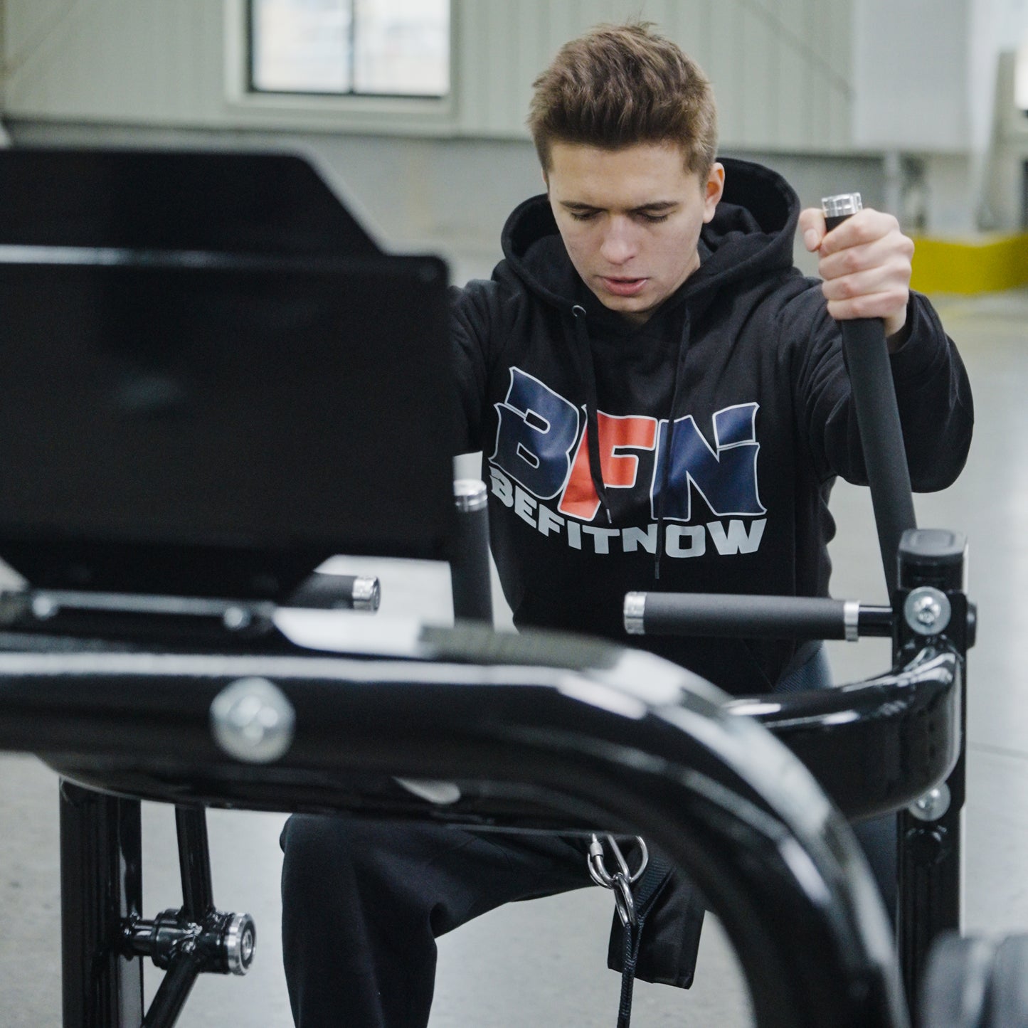 A young man wearing a BefitNow Canada hoodie uses the Commercial Belt Squat Trainer in a gym, focusing intently on his workout.