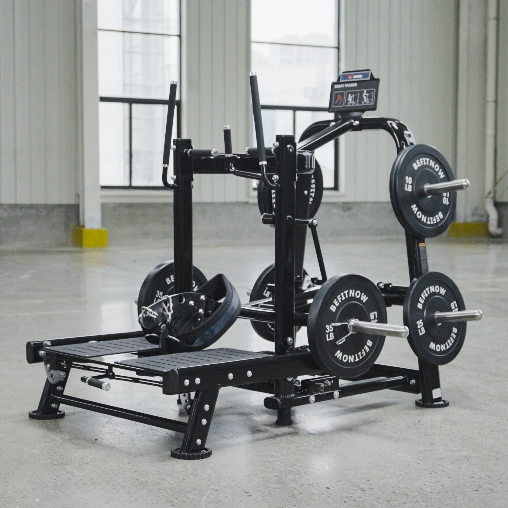 The BefitNow Canada Commercial Belt Squat Trainer, equipped with multiple weight plates, is set on a concrete floor in a spacious gym featuring large windows and white walls.