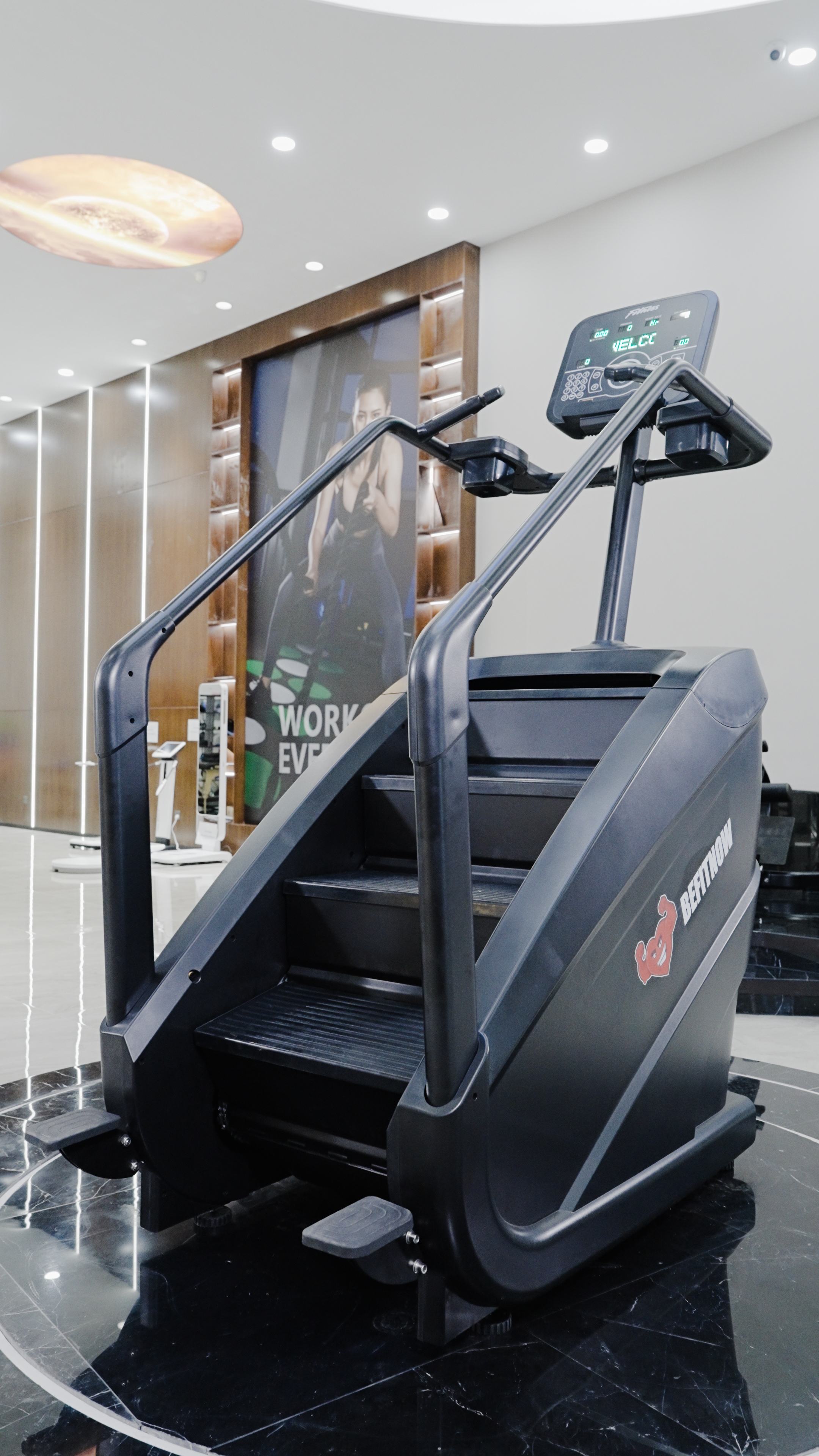 The BefitNow Canada Stair Climber 3000 - The Peak Punisher, a black commercial-grade machine, stands on a shiny black gym floor with wood wall panels and a workout poster in the background—enjoy joint-safe climbing every session.