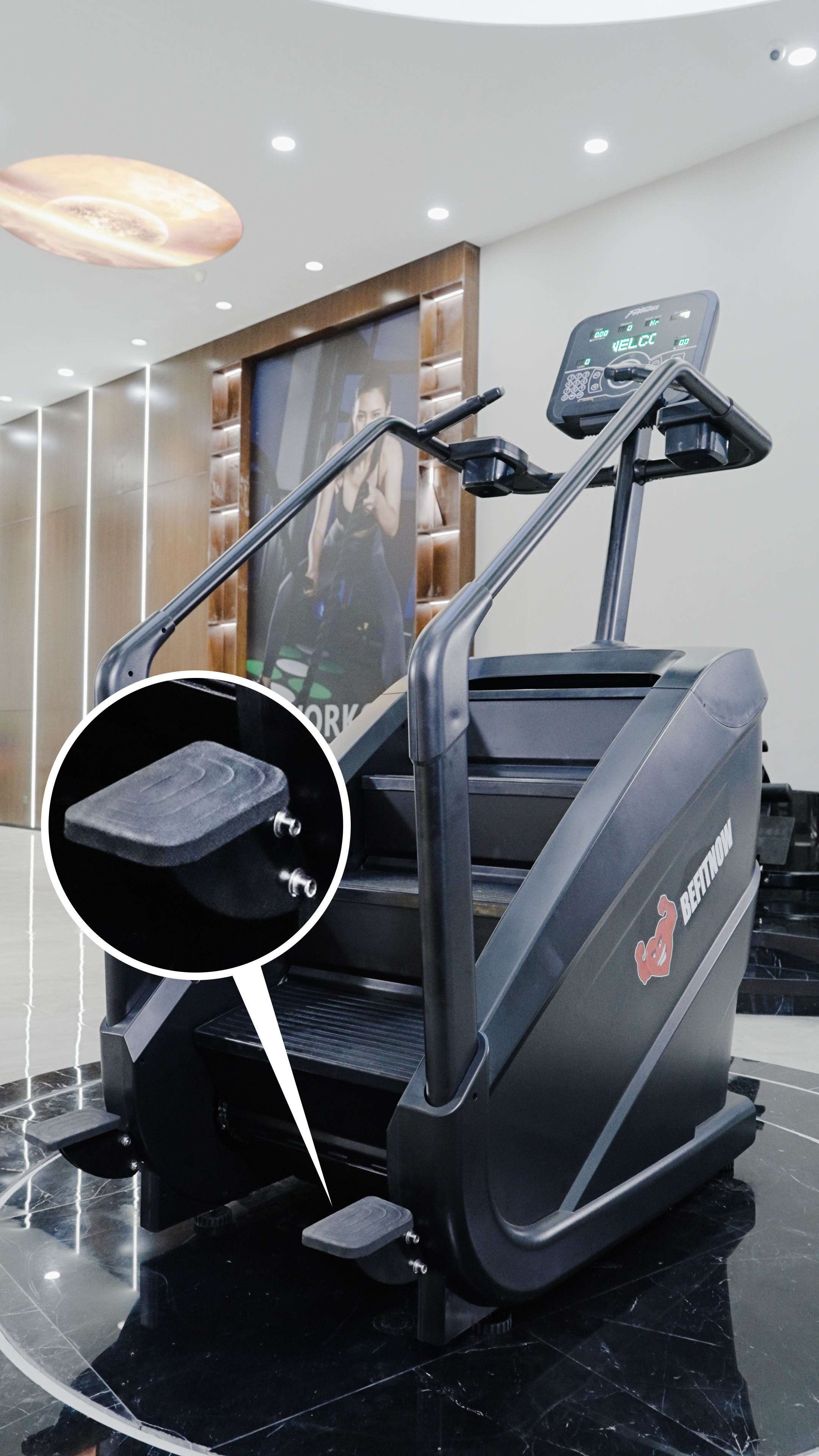 The BefitNow Canada Stair Climber 3000 - The Peak Punisher stands on a glossy black gym floor, with a close-up showcasing its joint-safe climbing pedal. A large workout poster decorates the background of the modern gym.