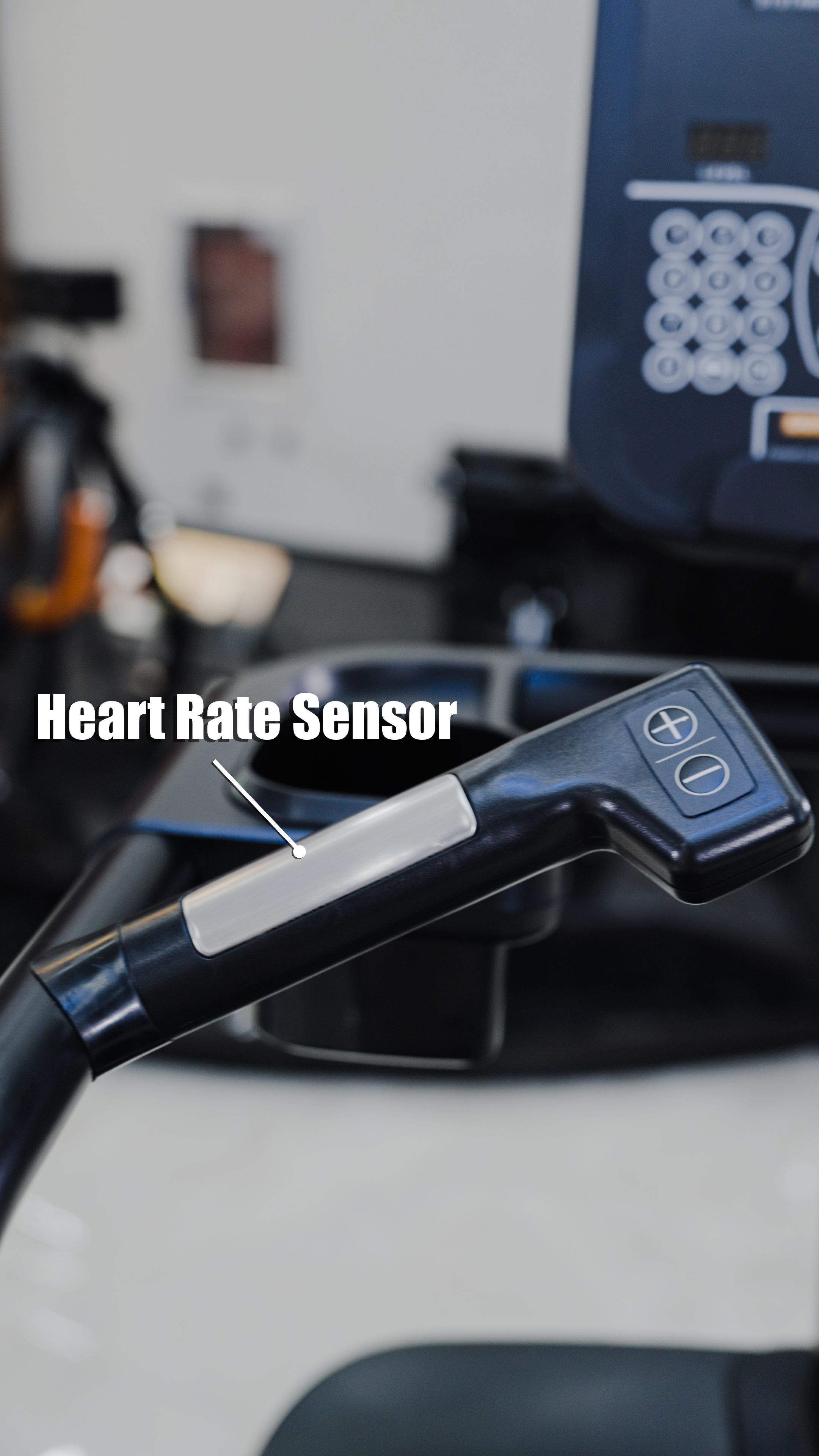 Close-up of a treadmill handle with a silver heart rate sensor and control buttons. In the background, a blurred BefitNow Canada Stair Climber 3000 – The Peak Punisher, featuring joint-safe climbing technology, is visible.