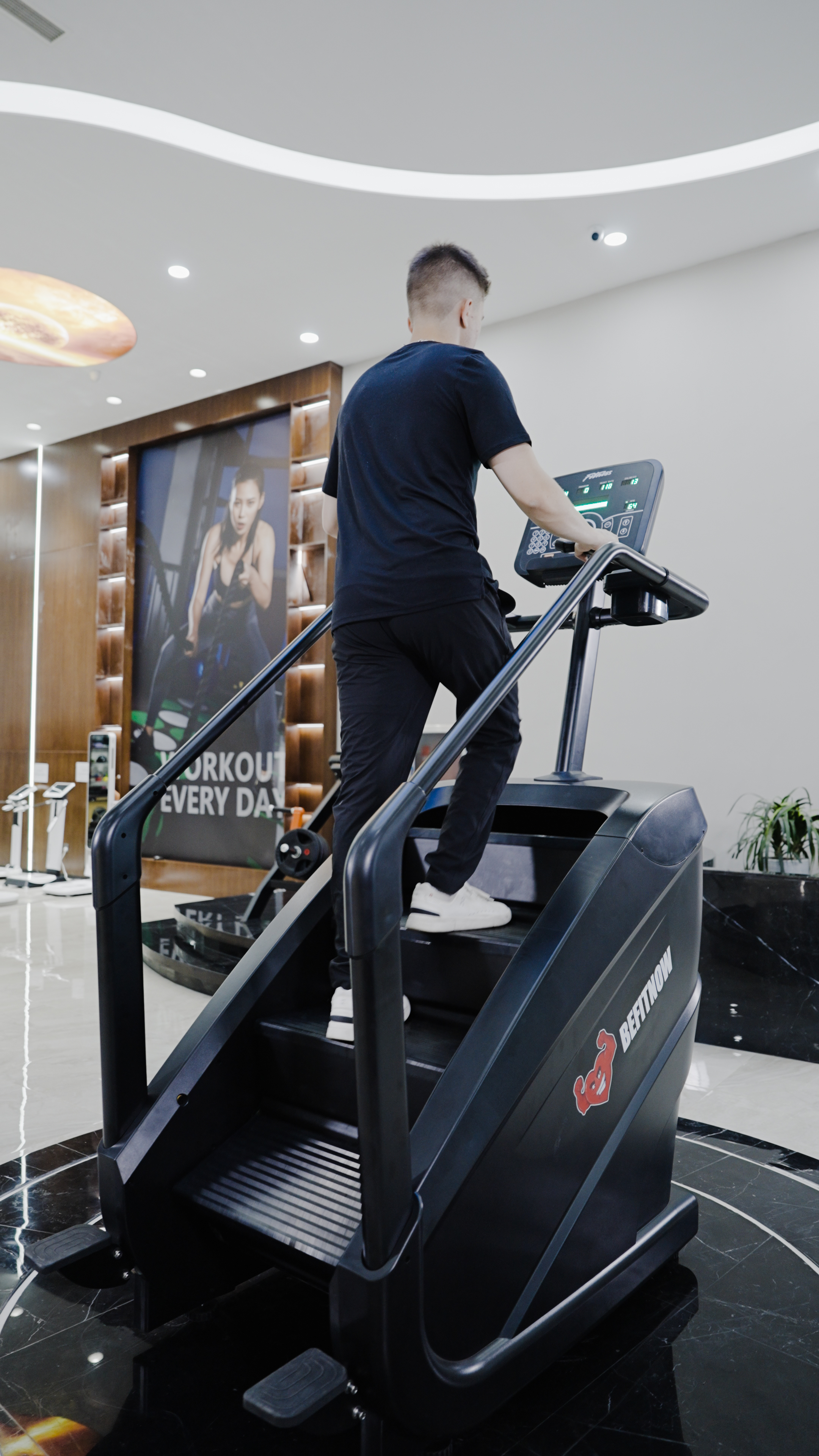 A man in black athletic wear uses the BefitNow Canada Stair Climber 3000 - The Peak Punisher, featuring joint-safe climbing technology, in a modern gym with a large workout poster of a woman visible on the wall.