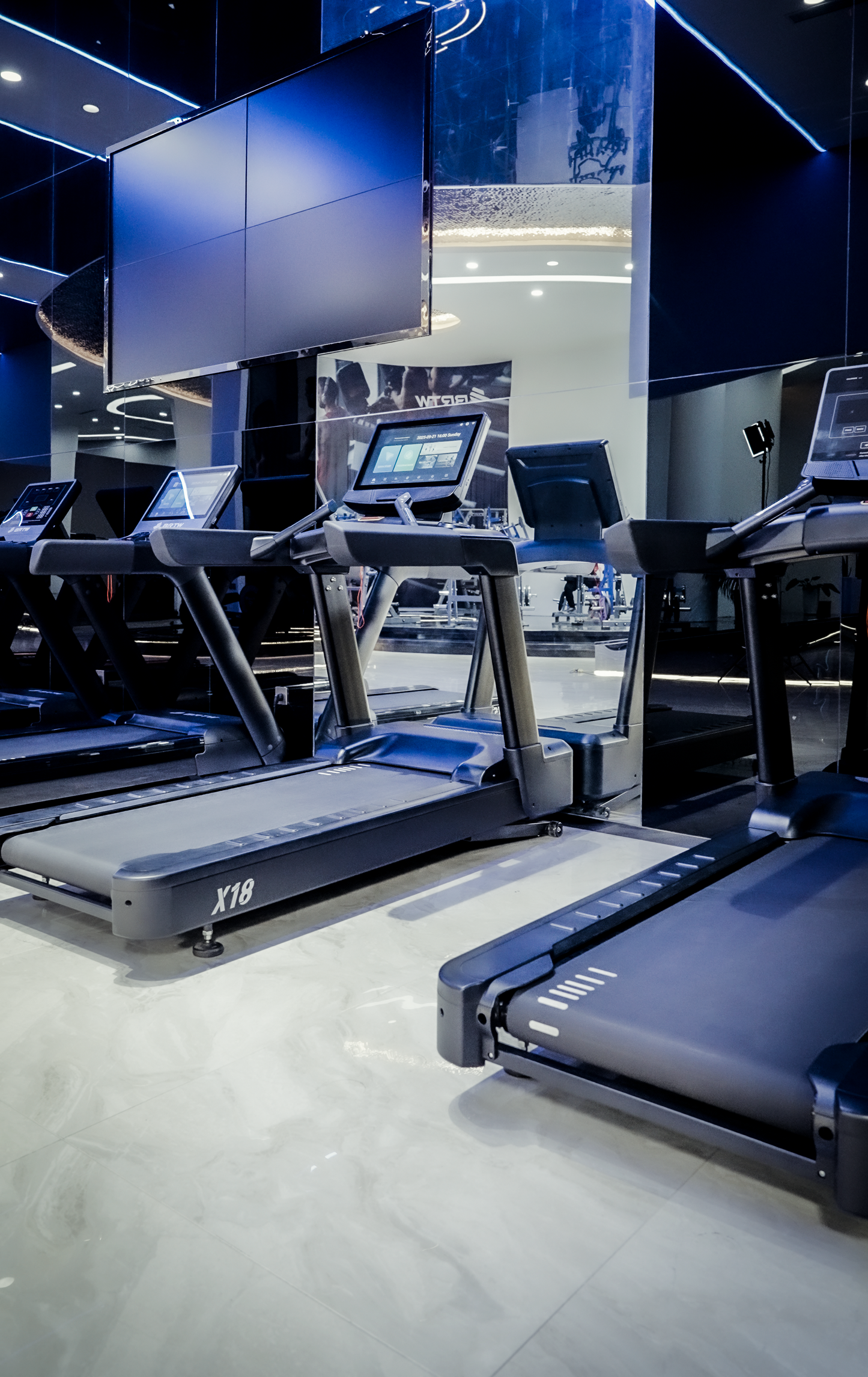 The NitroStep by BefitNow Canada treadmills, featuring oversized running belts and digital screens, are lined up in a sleek, brightly lit gym with glossy floors, mirrored walls, and large TV screens mounted above.