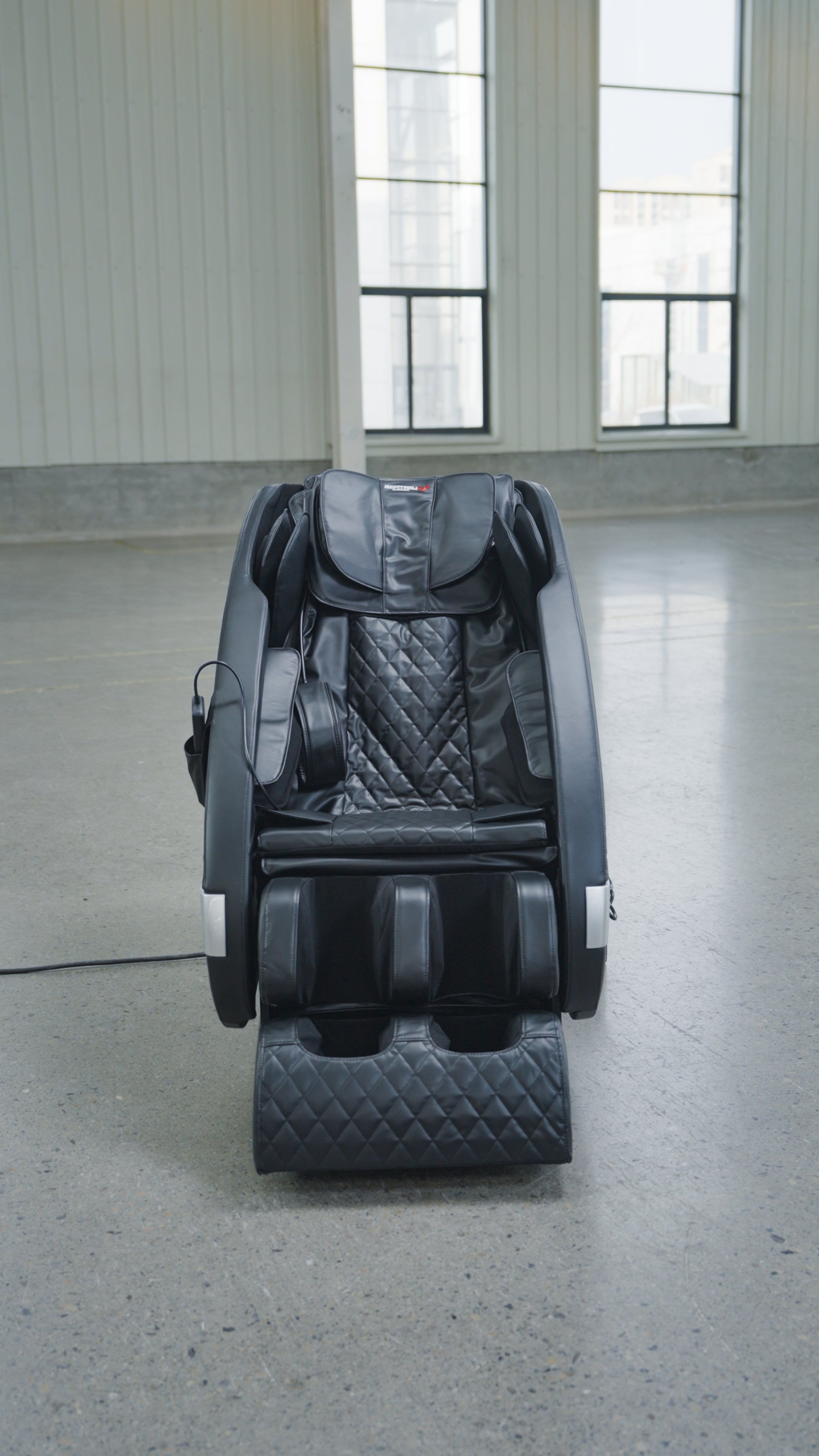 The BefitNow Canada Luxurious Zero Gravity Massage Chair in black, featuring quilted padding and a modern design, sits on a polished concrete floor in a spacious room with tall windows and white paneled walls.