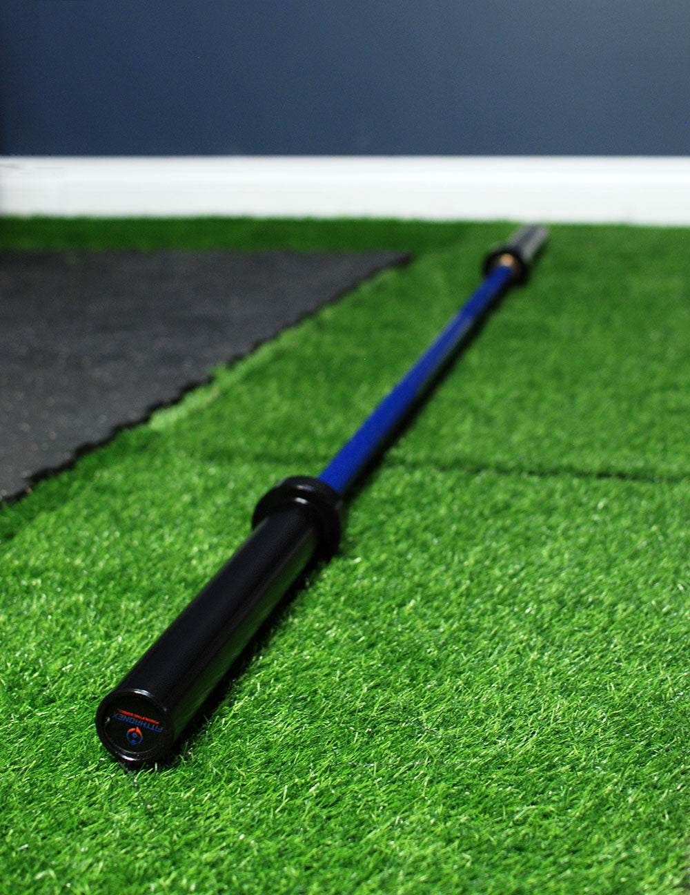 A Custom Barbells product from BefitNow Canada features black ends and a blue grip, resting on artificial green grass. It is surrounded by black matting in front of a dark blue wall, perfect for fitness enthusiasts.