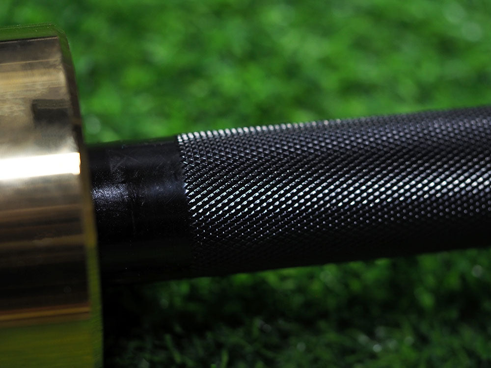 A close-up of a textured grip on a BefitNow Canada Custom Barbell handle transitions into a shiny, smooth section, set against a blurred green background resembling grass or turf.