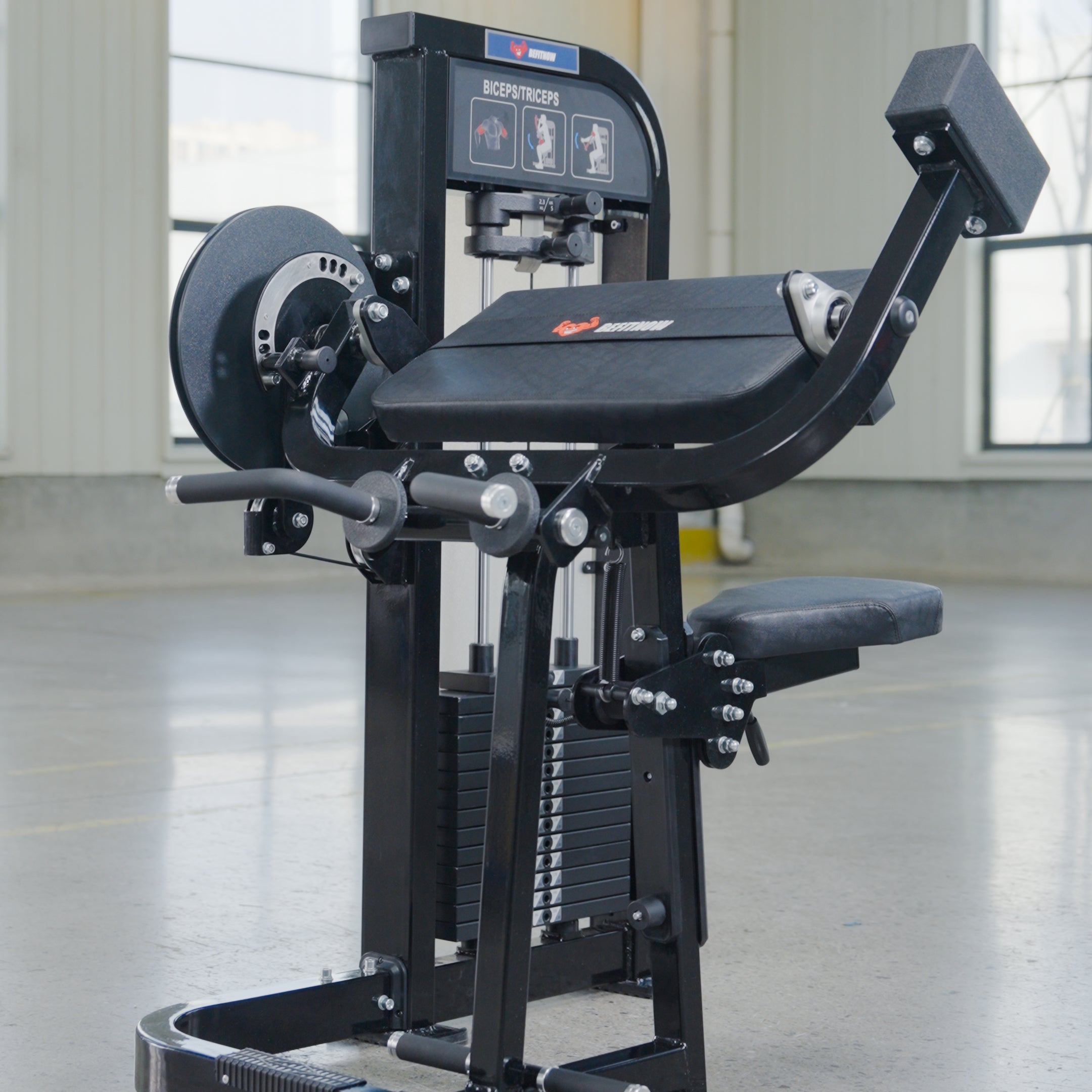 The BefitNow Canada Dual Function Biceps/Triceps machine with a black seat, padded supports, and weight plates stands on a smooth concrete floor beside a back extension unit in a bright gym with large windows.