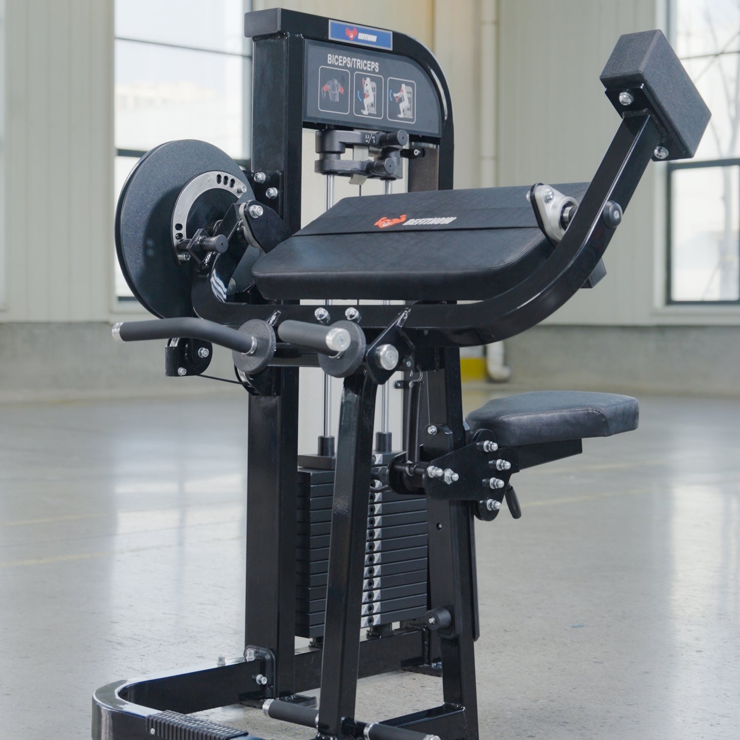 The BefitNow Canada Dual Function Biceps/Triceps machine with a black seat, padded supports, and weight plates stands on a smooth concrete floor beside a back extension unit in a bright gym with large windows.