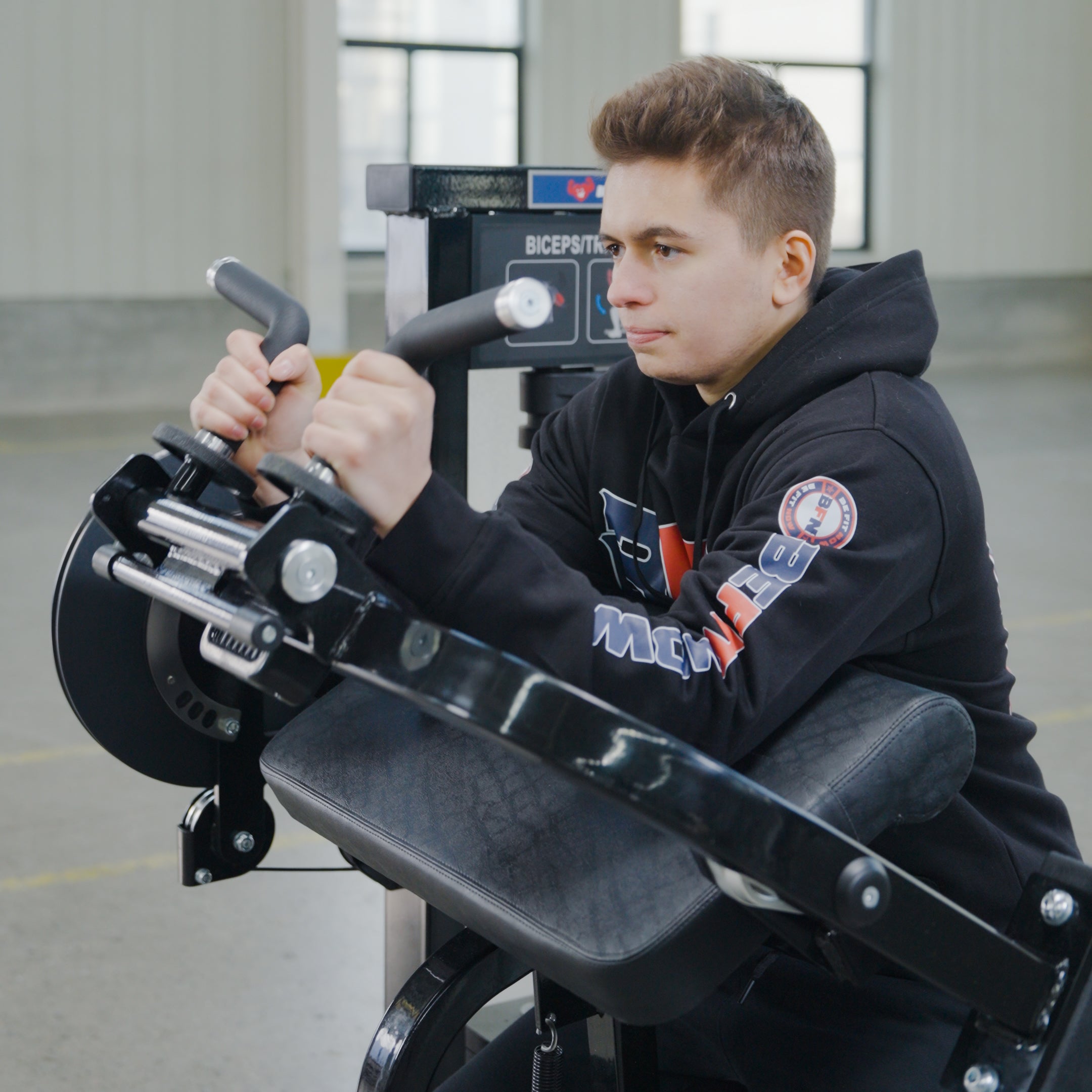 A young man in a black hoodie trains his arms on the BefitNow Canada Dual Function Biceps/Triceps machine, gripping its handles in an industrial-style gym.