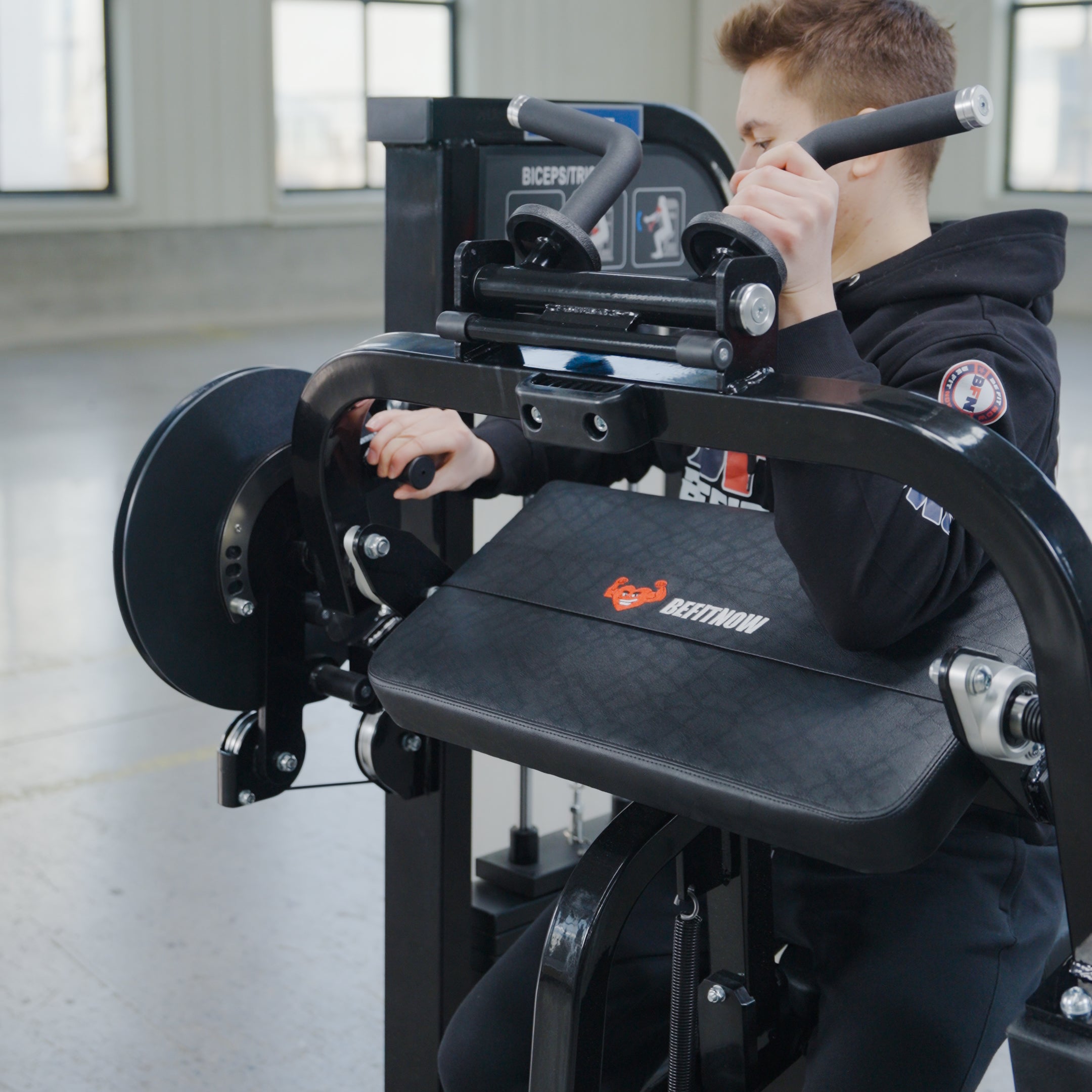 A person trains intensely on the BefitNow Canada Dual Function Biceps/Triceps machine in a gym, wearing a black hoodie with a sleeve logo. The adjustable resistance enhances focused arm workouts.