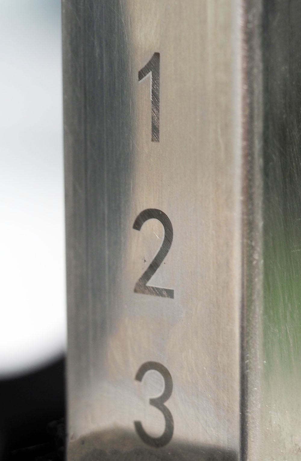 Close-up of an 11 gauge steel column on the TM87 Fully Adjustable Commercial Bench by BefitNow Canada, engraved with numbers 1, 2, and 3 in a vertical stack. The smooth, reflective surface partially reveals a blurred background, adding intrigue.