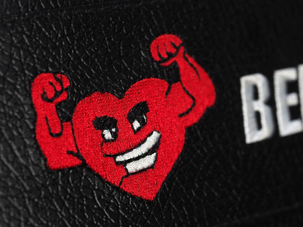 A cartoon heart with a grin, flexed arms, bold eyebrows, and eyes is embroidered on a black surface of your BefitNow Canada TM87 Fully Adjustable Commercial Bench. The letters BE are partially visible on the premium textured foam pad.