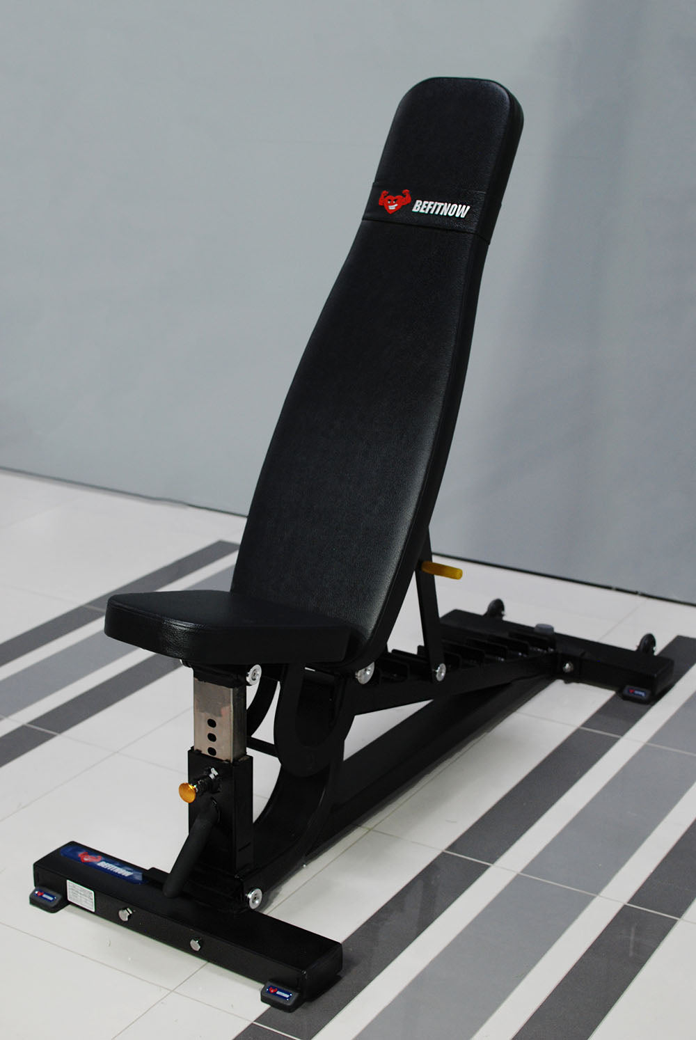 The BefitNow Canada TM87 Fully Adjustable Commercial Bench, perfect for gyms, comes in black and gray with a branded padded backrest and seat. It complements the light gray wall and tiled floor beautifully.